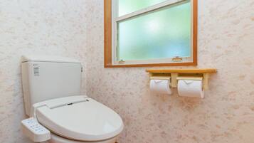 Sanitário com painel de controle, uma janela acima e rolo de papel higiênico na parede.