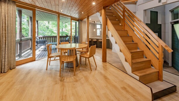 Une pièce au plancher de bois, avec une table ronde et des chaises, un escalier et de grandes fenêtres.