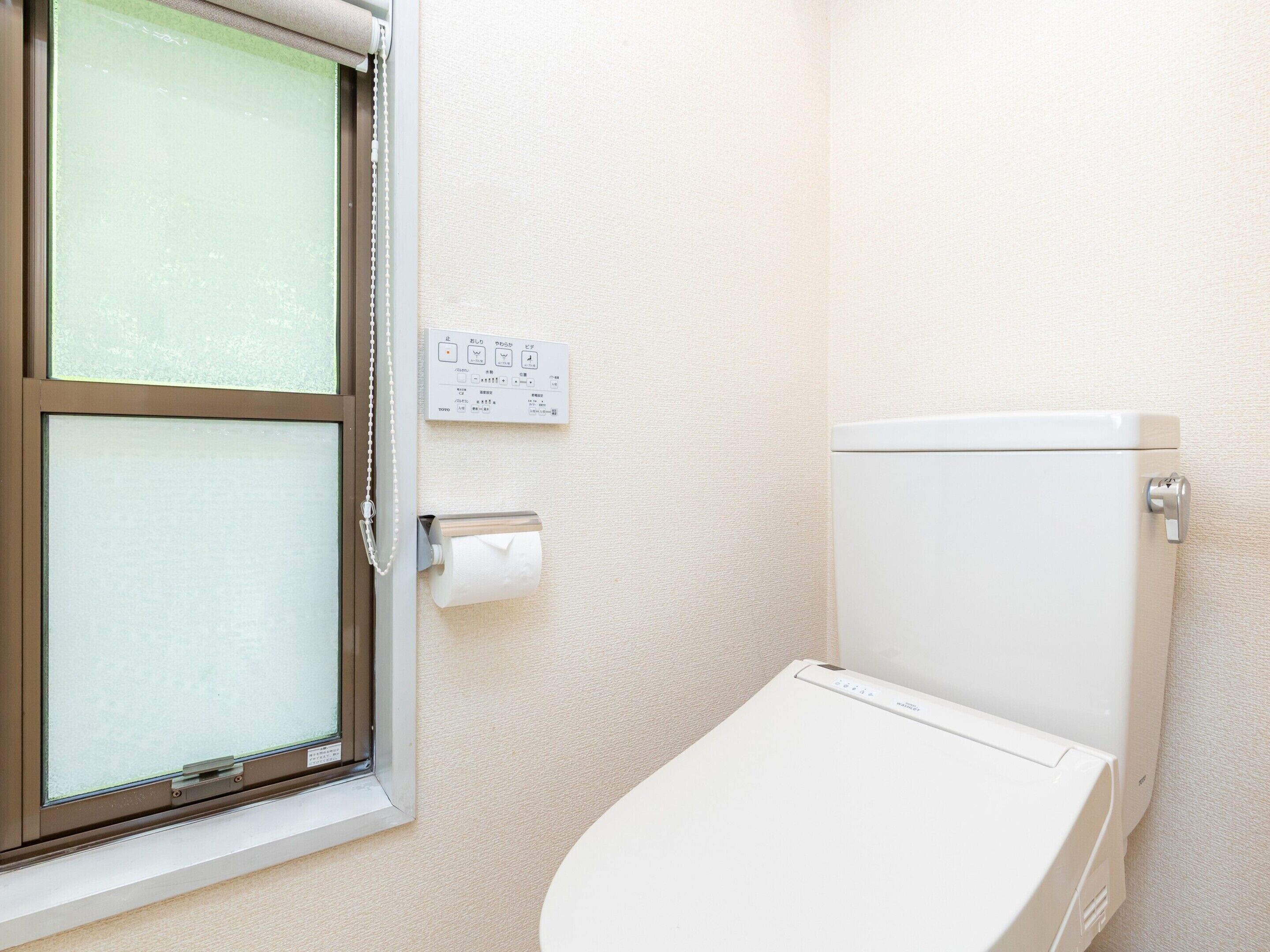 A bathroom with a toilet, a window with blinds, and a wall-mounted flush handle.