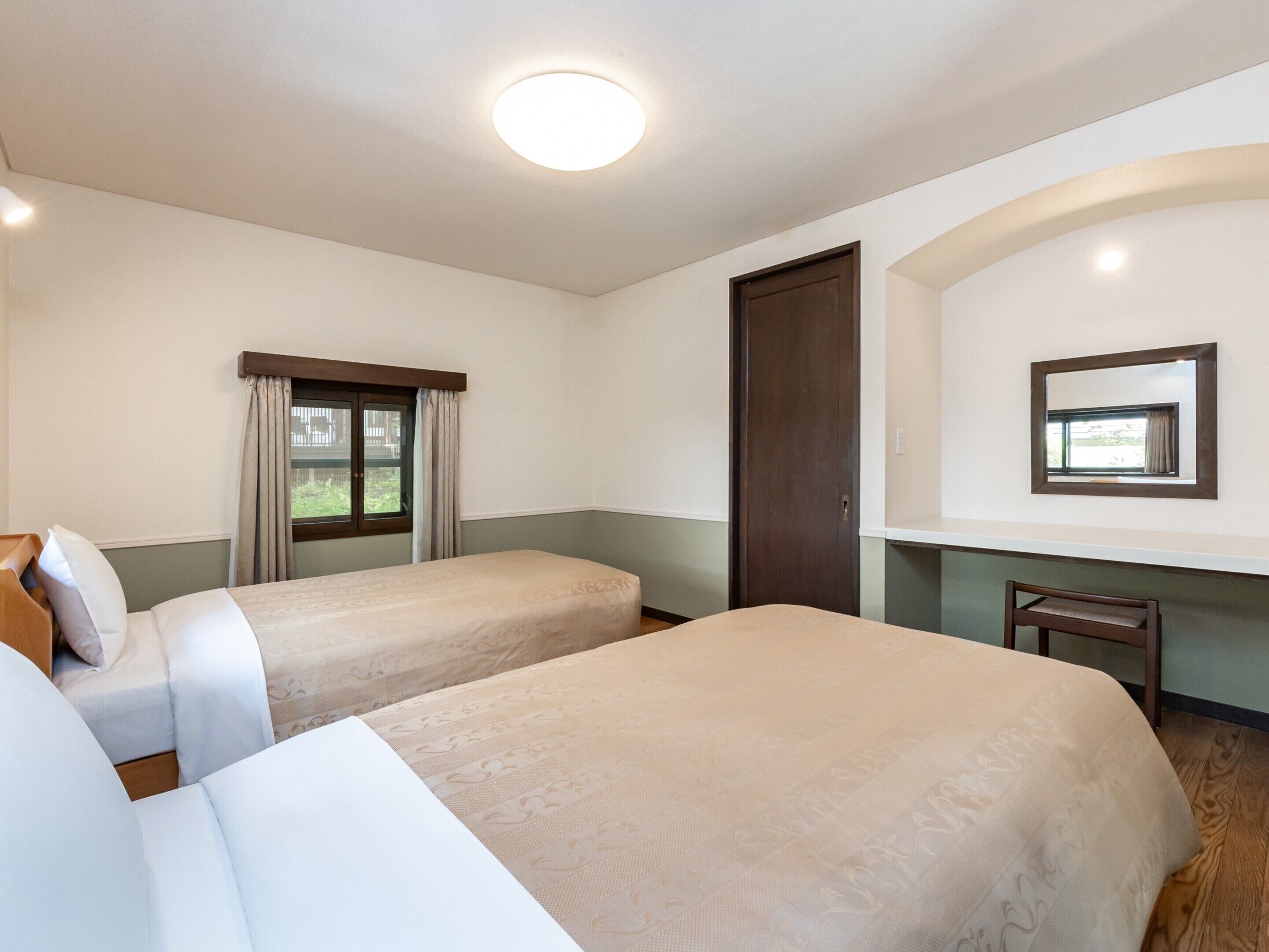 A hotel room with two beds, a window with curtains, a door, a mirror, and a small desk.