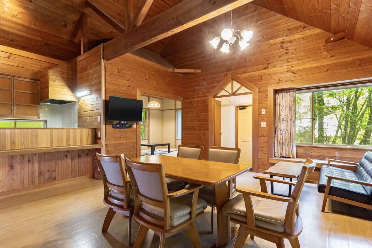 Interno di una baita in legno con zona pranzo, televisore montato a parete e finestra vista alberi.