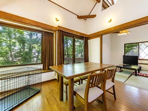 Una habitación con una mesa de comedor de madera, sillas, un televisor y vistas a árboles a través de ventanas amplias.