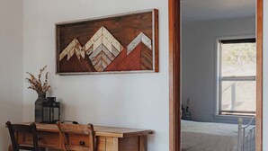 A room with a wooden dining table, chairs, a wall-mounted painting, and a bed in the adjacent room.