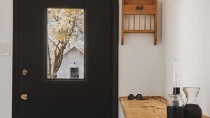 Une porte noire avec une fenêtre, une étagère en bois ornée d’une découpe en forme de cœur, un tabouret et une table sur laquelle se trouvent un récipient en verre et un pichet en métal.