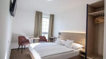 Quarto de hotel com cama, escrivaninha, duas cadeiras, guarda-roupa e televisão na parede.