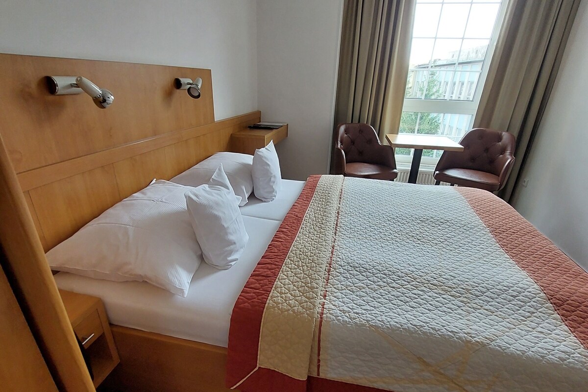 Ein Hotelzimmer mit einem hölzernen Kopfteil, einem Bett mit weißer Bettwäsche, zwei Stühlen, einem kleinen Tisch und einem Fenster mit Vorhängen.