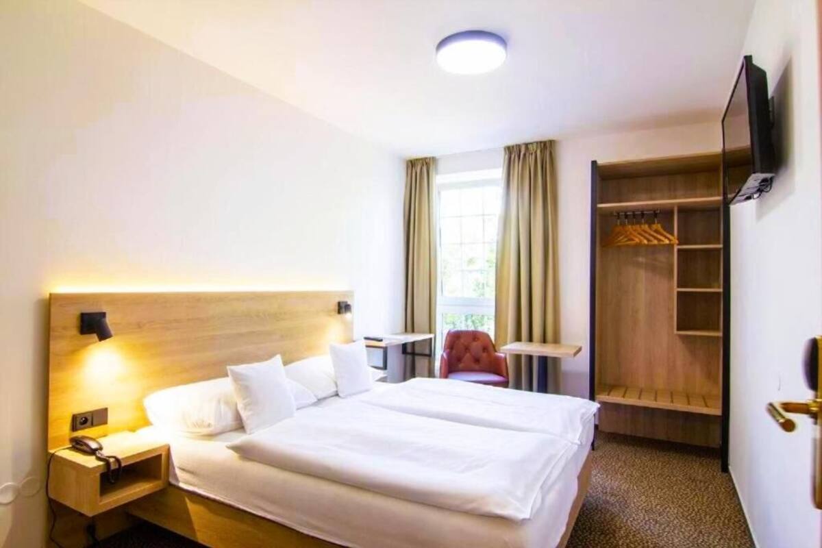 A hotel room with a large bed, bedside table with a lamp, a small desk, a chair, a wardrobe, and a TV mounted on the wall.