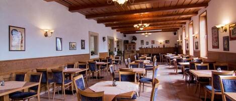 A spacious restaurant with wooden tables and chairs, high ceilings with exposed beams, and wall-mounted lighting.