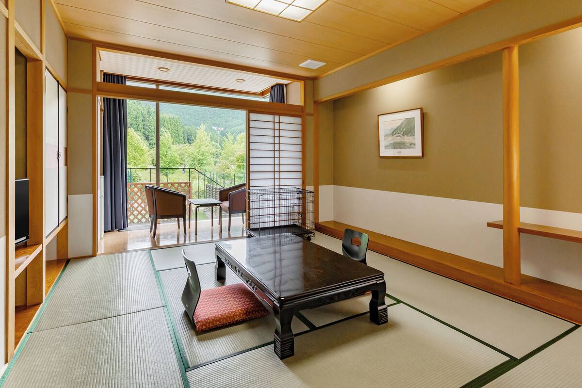 A traditional Japanese-style room with tatami flooring, a low table, a cushioned bench, and a sliding door leading to a balcony with a view of greenery.
