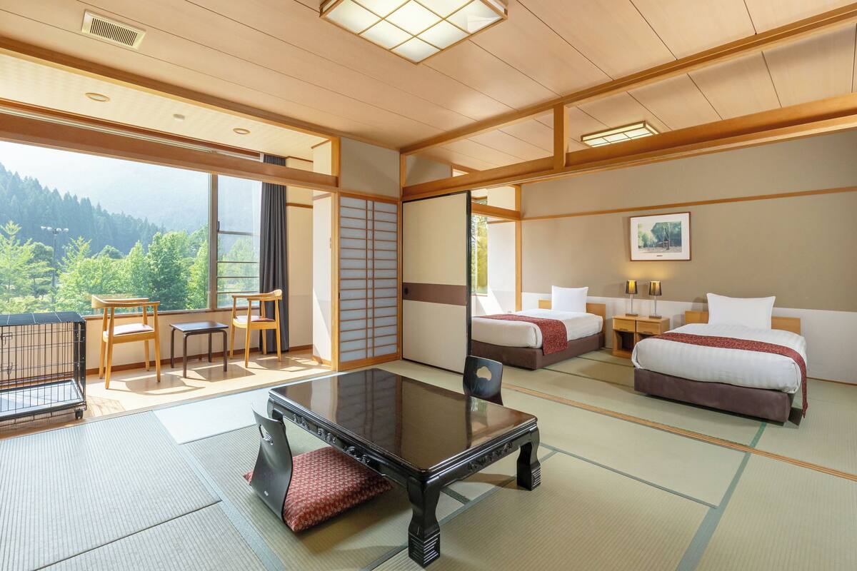A traditional Japanese-style room with tatami flooring, a low table, two beds, and a view of a forested area.