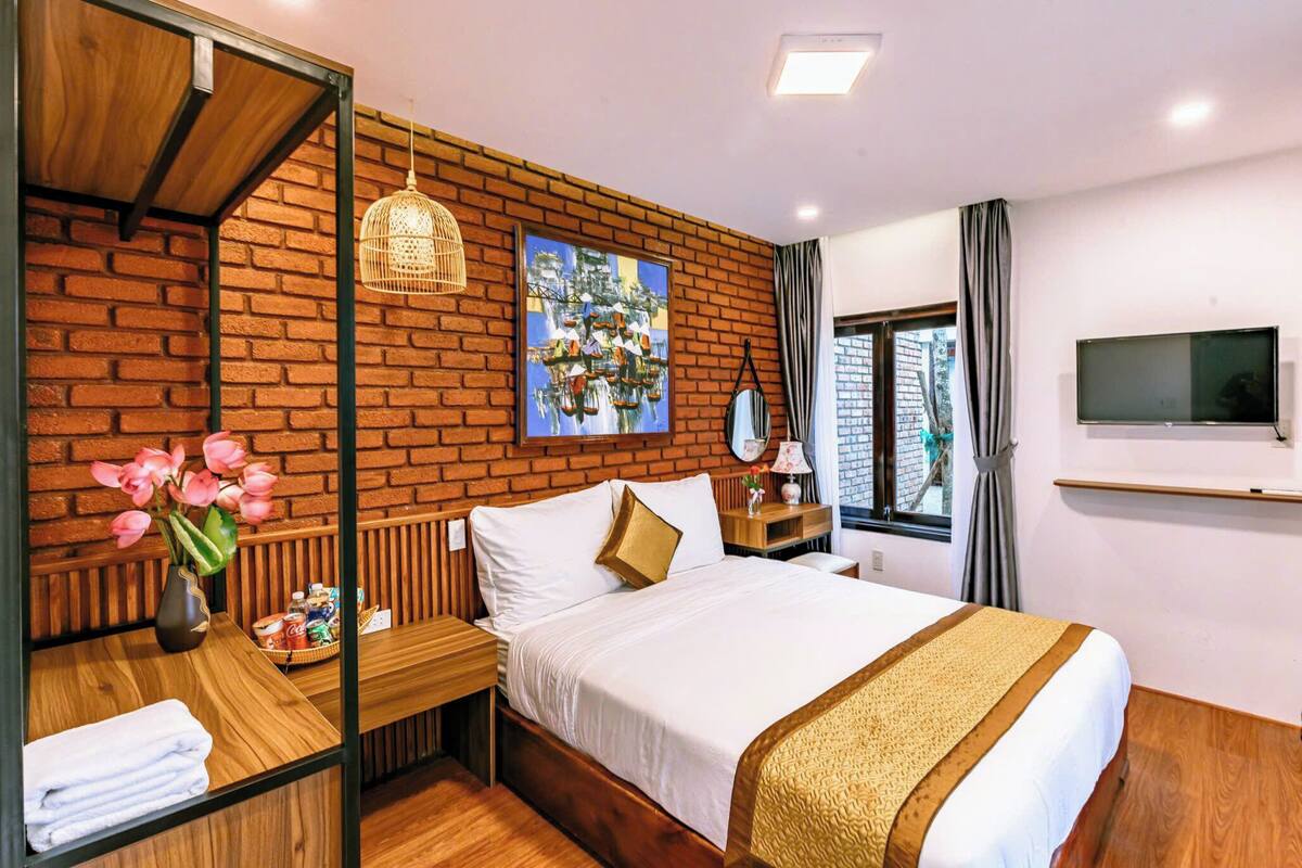 A hotel room with a brick wall, a bed with white linens, a wooden nightstand, a vase with flowers, a television on the wall, and a window with curtains.