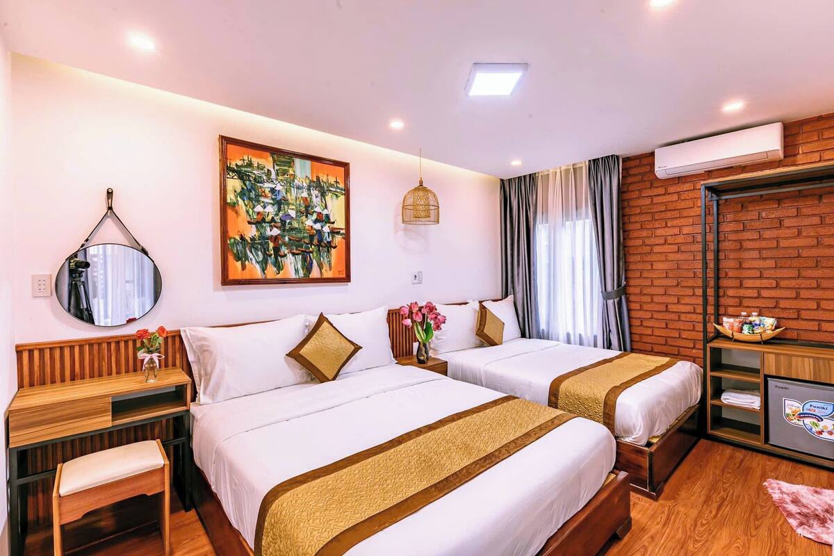 A hotel room with two beds, a desk, a mirror, a painting, a hanging light fixture, an air conditioning unit, and a television.