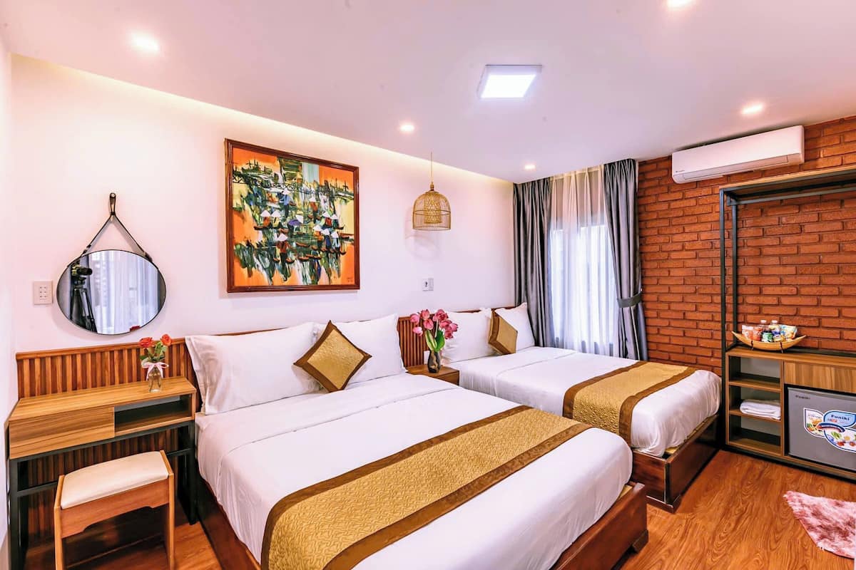 A hotel room with two beds, a desk, a mirror, a painting, a hanging light fixture, an air conditioning unit, and a television.