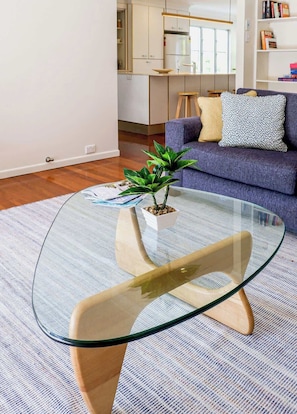 A modern living room with a glass coffee table, a sofa with cushions, a small plant, and a kitchen in the background.
