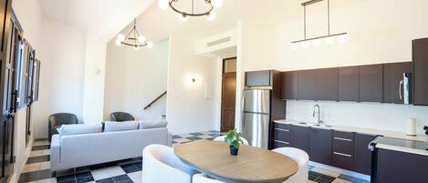 A modern kitchen with a refrigerator, stove, and dark cabinets. There is a dining area with a table and chairs, and a sofa in the background.