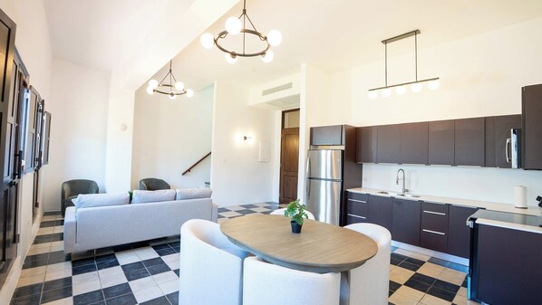 A modern kitchen with a refrigerator, stove, and dark cabinets. There is a dining area with a table and chairs, and a sofa in the background.