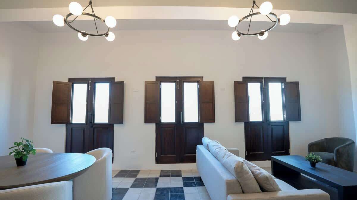 A modern living room with a sofa, a coffee table, and a dining table. There are three large windows with dark frames and a chandelier hanging from the ceiling.