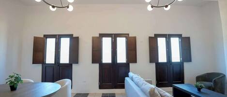 A modern living room with a sofa, a coffee table, and a dining table. There are three large windows with dark frames and a chandelier hanging from the ceiling.