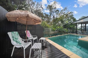 Ein Poolbereich mit Holzterrasse, einem Strohdachschirm, weißen Liegestühlen und einem Pool.