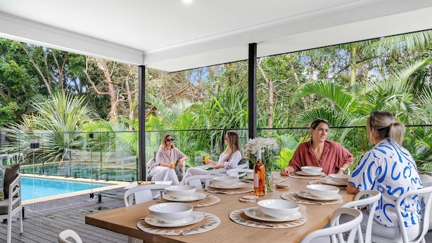 Un comedor con una mesa de madera dispuesta para seis personas, rodeado de sillas blancas, con vistas a una piscina y abundante vegetación.