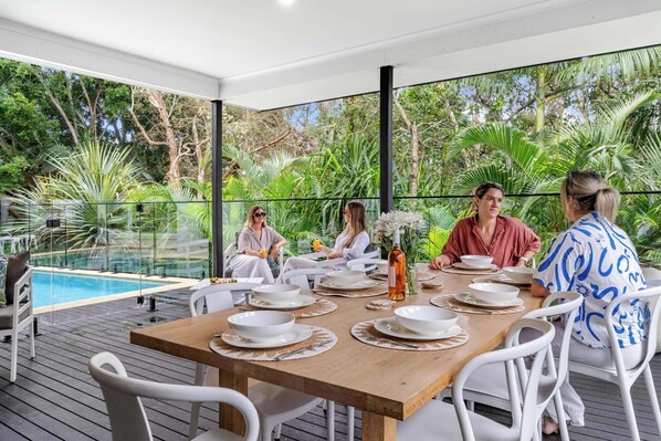 Un comedor con una mesa de madera dispuesta para seis personas, rodeado de sillas blancas, con vistas a una piscina y abundante vegetación.