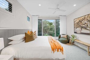 A modern bedroom with a large bed, a ceiling fan, a window with a view of greenery, and a wall-mounted artwork.