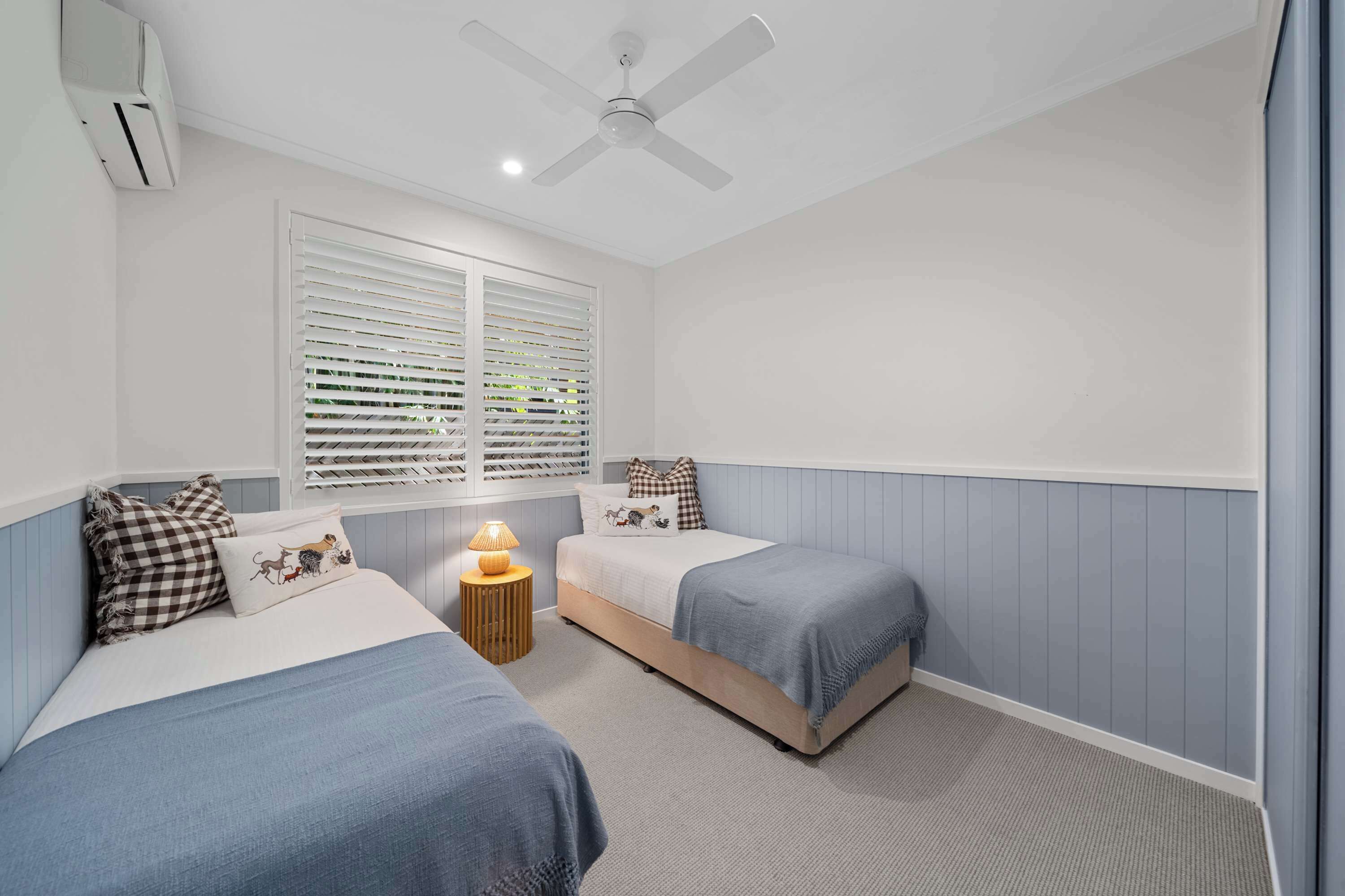 Une chambre avec deux lits, un ventilateur de plafond, une fenêtre avec des stores et une lampe de chevet.