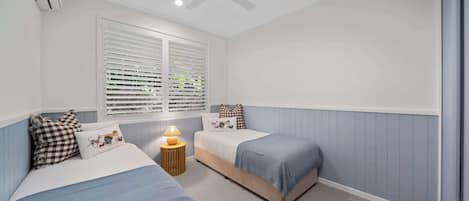 Un dormitorio con dos camas, ventilador de techo, ventana con persianas y una lámpara de noche.