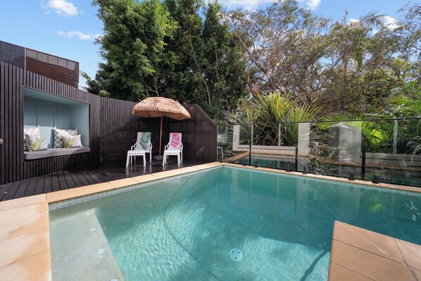 A rectangular swimming pool with a wooden deck, two white chairs, a thatched umbrella, and a built-in seating area with cushions.