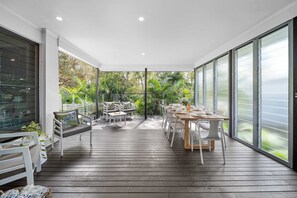 A modern outdoor dining area with a wooden deck, white furniture, and large glass doors leading to a lush garden.