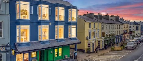 Una hilera de edificios con escaparates, incluyendo un comercio con fachada verde y un edificio azul con amplias ventanas.