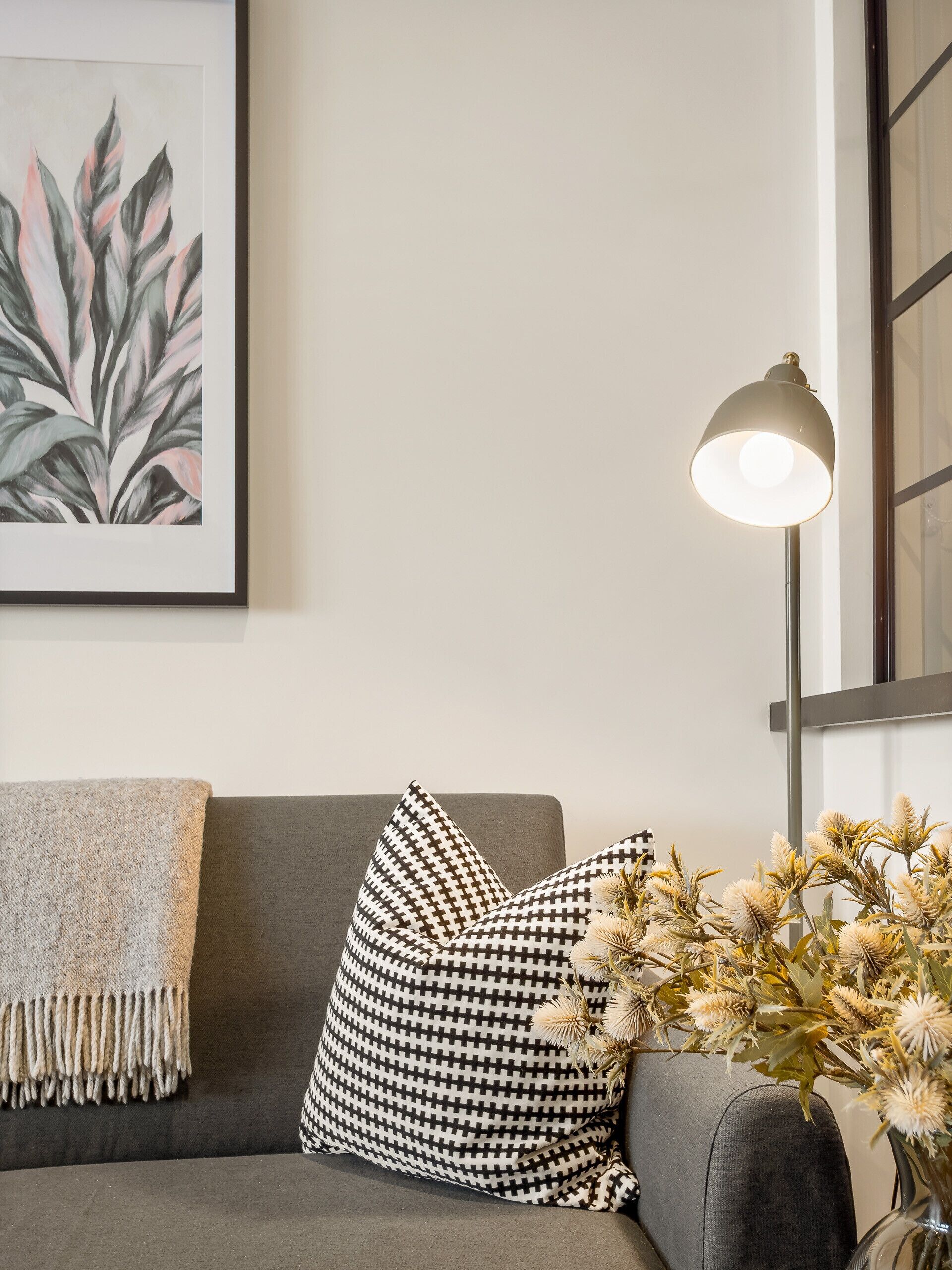 Un salon moderne avec un canapé gris, des coussins à motifs, une estampe botanique encadrée, une lampe sur pied et un vase contenant des fleurs séchées.