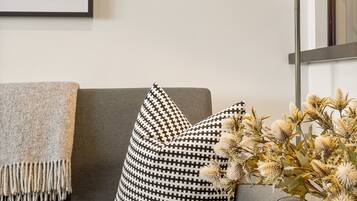 Un salon moderne avec un canapé gris, des coussins à motifs, une estampe botanique encadrée, une lampe sur pied et un vase contenant des fleurs séchées.
