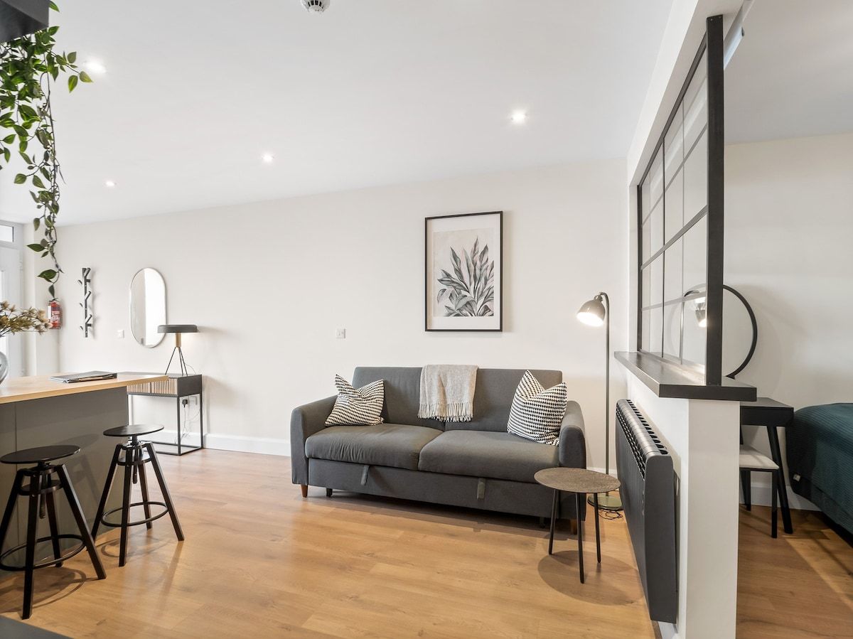 Ein modernes Wohnzimmer mit einem grauen Sofa, zwei schwarzen Barhocker, einem kleinen runden Tisch und einer Heizung.