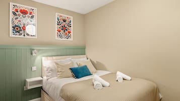 Un letto rifatto con lenzuola bianche e un cuscino blu, un comodino con una lampada, e due opere d'arte floreali appese al muro.
