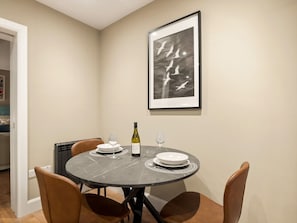 A dining area with a round table, four chairs, two wine glasses, a bottle of wine, and a framed picture on the wall.