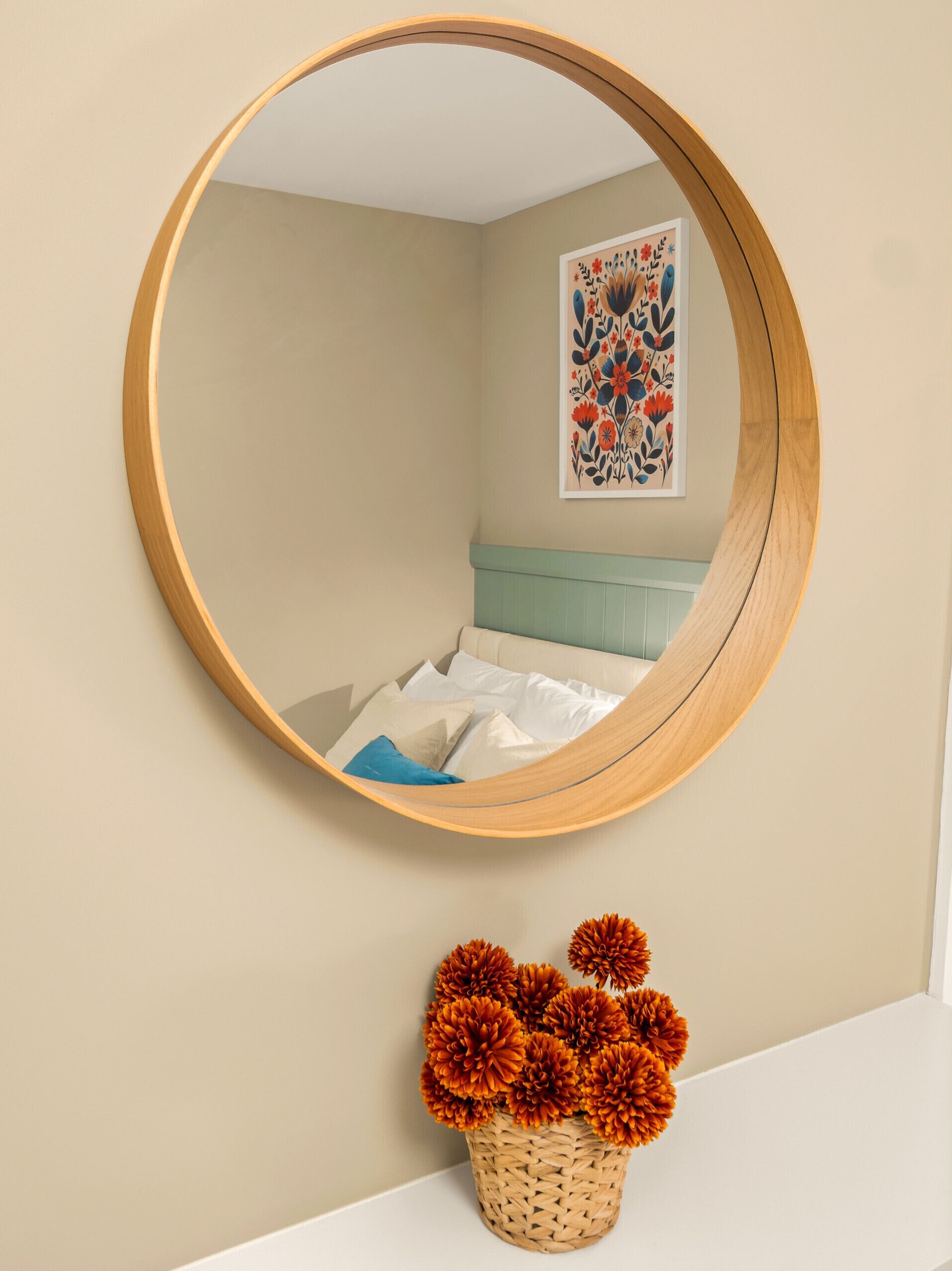 Et rundt spejl på væggen, en seng med et blåt sengegærde, et blomsterkunstværk og en kurv med orange blomster.