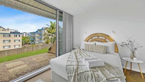 A bedroom with a large bed, a wooden headboard, a small bedside table, and a view of a balcony with a cityscape.