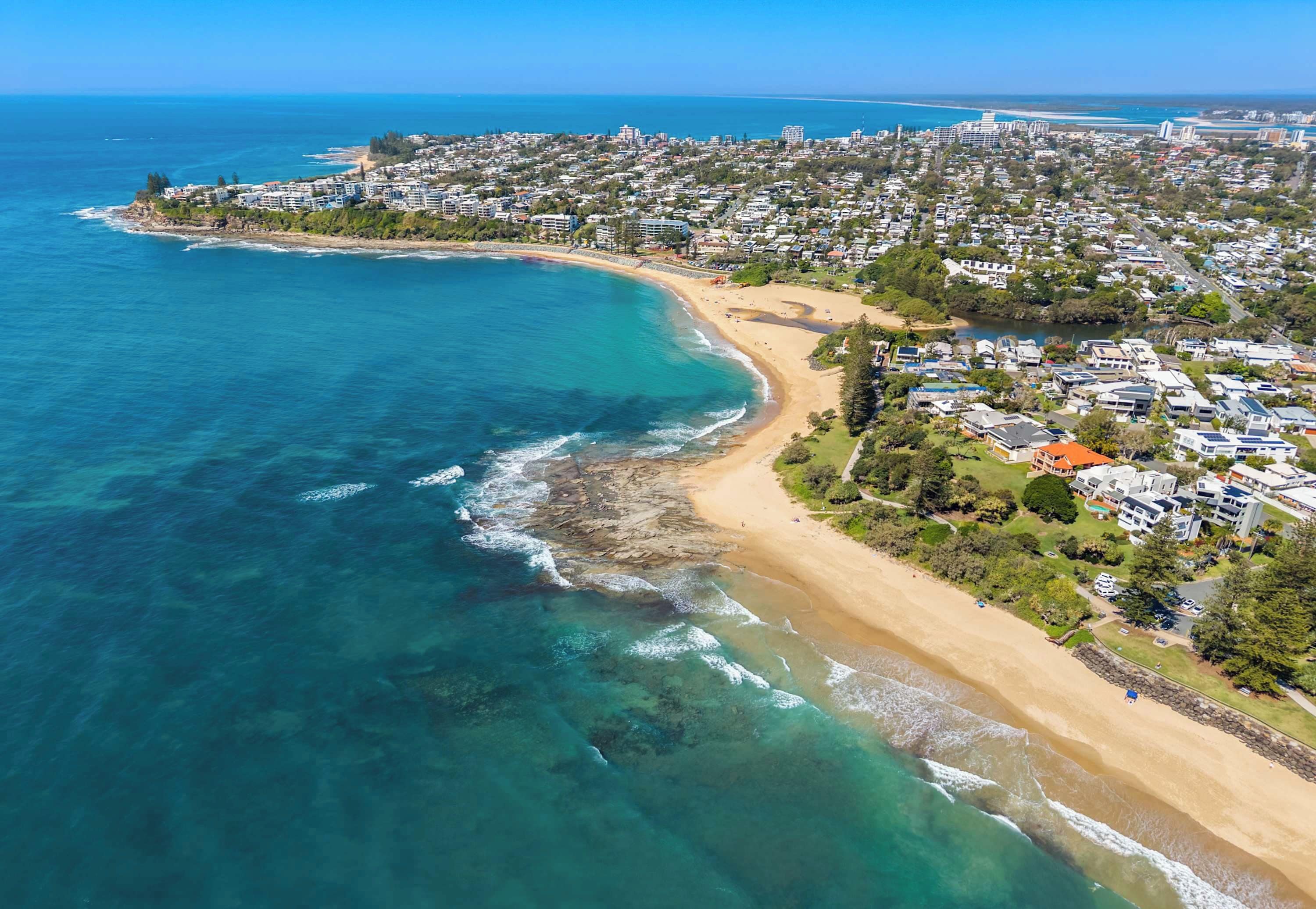 An aerial view of a coastal area with a beach, residential buildings, and a clear blue sky.