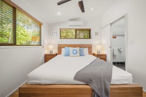 A bedroom with a bed, bedside tables, a ceiling fan, an air conditioning unit, a window with blinds, and a bathroom with a shower. - Delightful Dicky Beach Holiday House (Caloundra Region)