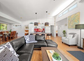 A modern living room with a black leather sofa, a dining area with wooden chairs, a kitchen with white cabinets, and a large window providing natural light. - Delightful Dicky Beach Holiday House (Caloundra Region)