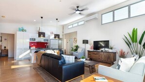 Uma sala de estar moderna com sofá, mesa de centro e TV. Cozinha visível ao fundo, com eletrodoméstico vermelho.