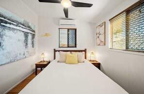 A bedroom with a bed, two bedside tables, a ceiling fan, an air conditioning unit, and a window with blinds. - Delightful Dicky Beach Holiday House (Caloundra Region)