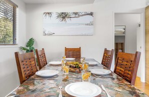 A dining area with a table set for four, surrounded by wooden chairs, with a tropical-themed painting on the wall and a window with blinds. - Delightful Dicky Beach Holiday House (Caloundra Region)
