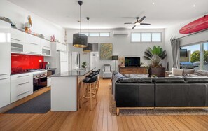 A modern kitchen with a red backsplash, a dining area with bar stools, a black leather sofa, and a large window with a view of the outdoors. - Delightful Dicky Beach Holiday House (Caloundra Region)