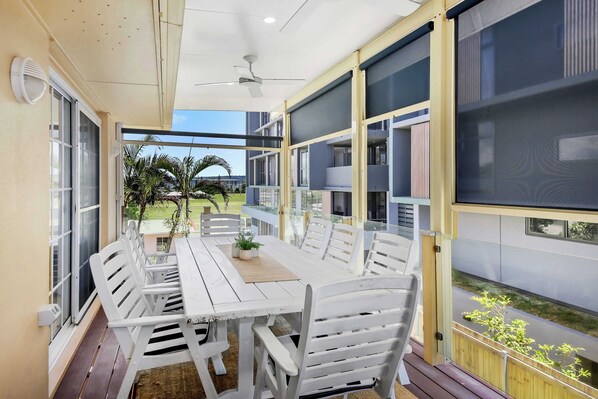 A modern outdoor dining area with a white table and chairs, large windows, and a view of the surrounding landscape.