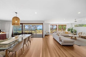 A spacious living area with a glass dining table, a sofa, and a coffee table. There are large windows offering a view of the outdoors.