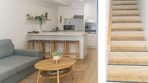 A modern kitchen with white cabinets, a microwave, and a stove. There is a dining area with a wooden table and a gray sofa. A staircase is visible to the right. - Escape to Coolum - Vibrant Beachside Lifestyle (Coolum Region)