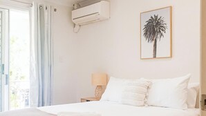 A bedroom with a bed, a nightstand, a lamp, an air conditioning unit, and a framed picture on the wall. - Escape to Coolum - Vibrant Beachside Lifestyle (Coolum Region)