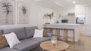 A modern living room with a grey sofa, a round wooden coffee table, and a kitchen area with white cabinets and a microwave. - Escape to Coolum - Vibrant Beachside Lifestyle (Coolum Region)